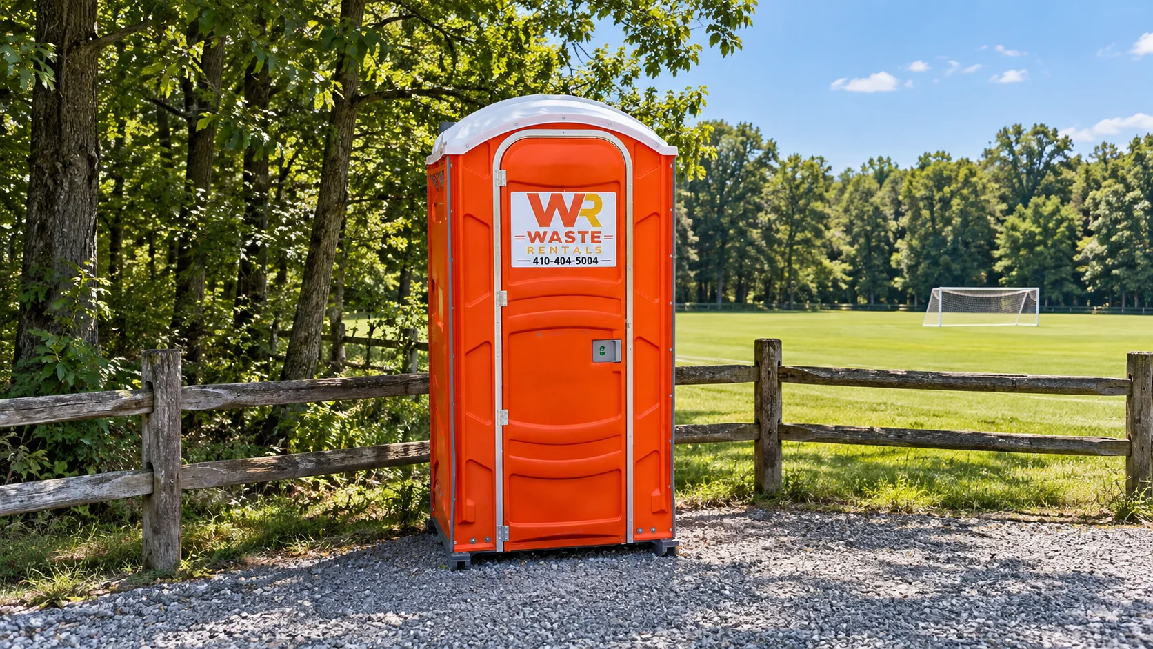 Orange Waste Rentals portable toilet on gravel beside a wooded sports field
