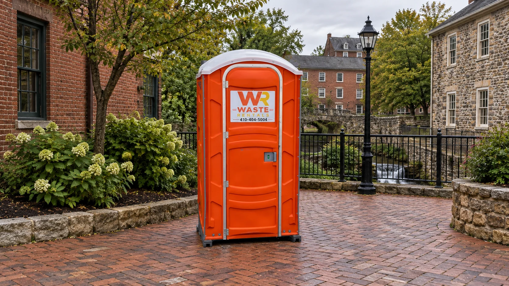 Orange Waste Rentals portable toilet in a brick courtyard with historic mill town buildings
