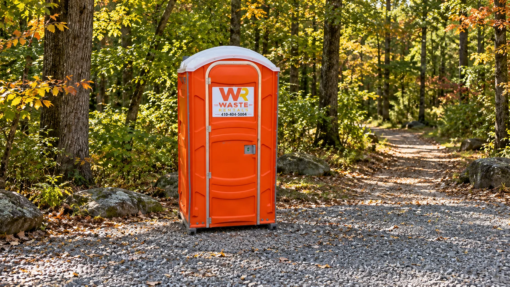 Orange Waste Rentals portable toilet on a level gravel clearing beside an autumn woodland trail