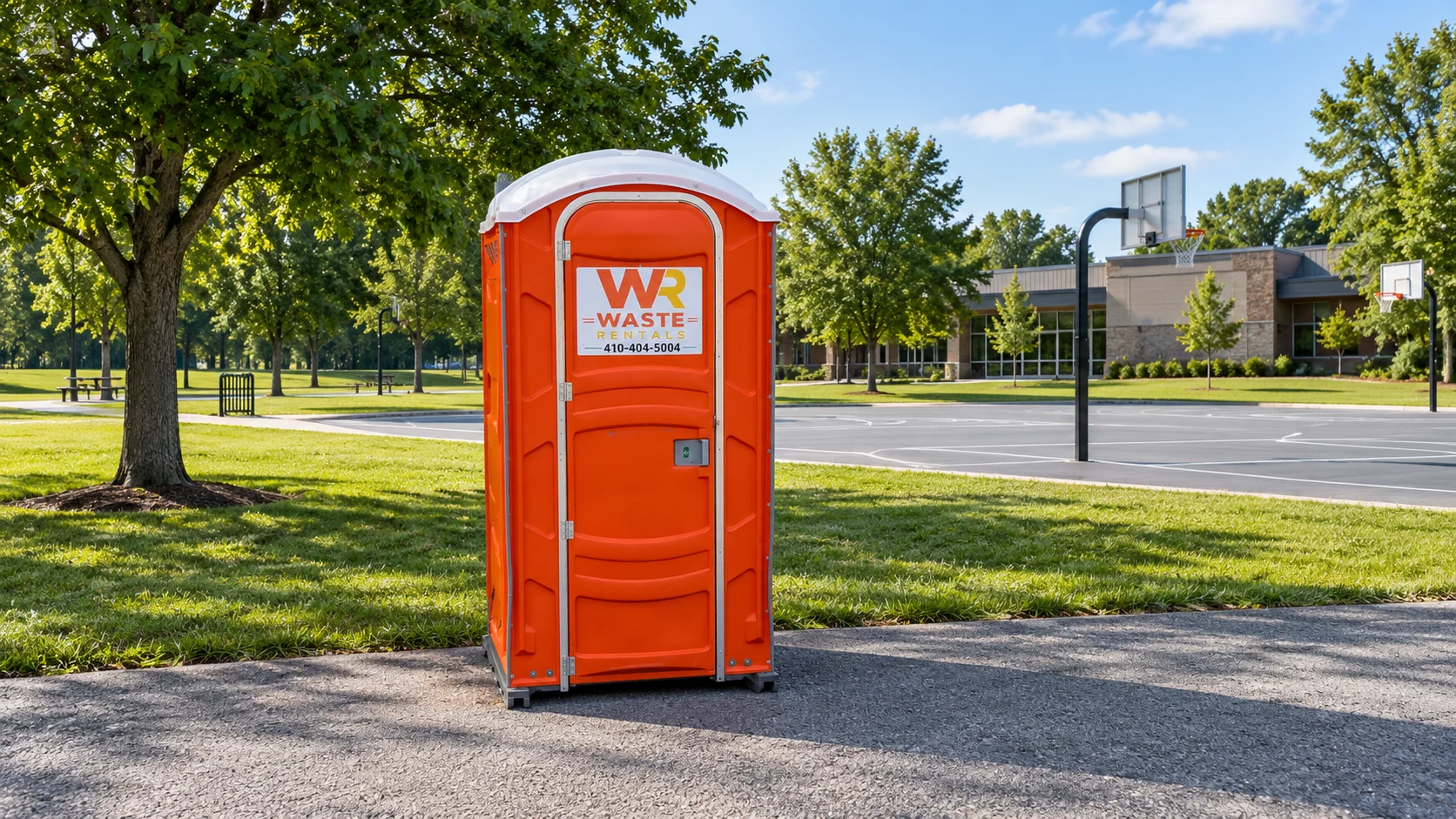 Orange Waste Rentals portable toilet beside a recreation lawn with courts in the distance