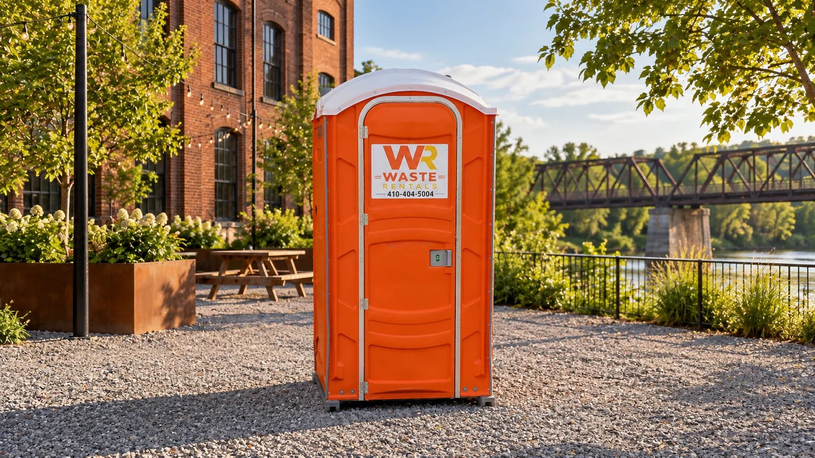 Orange Waste Rentals portable toilet beside a brick mill style event space with a distant iron bridge
